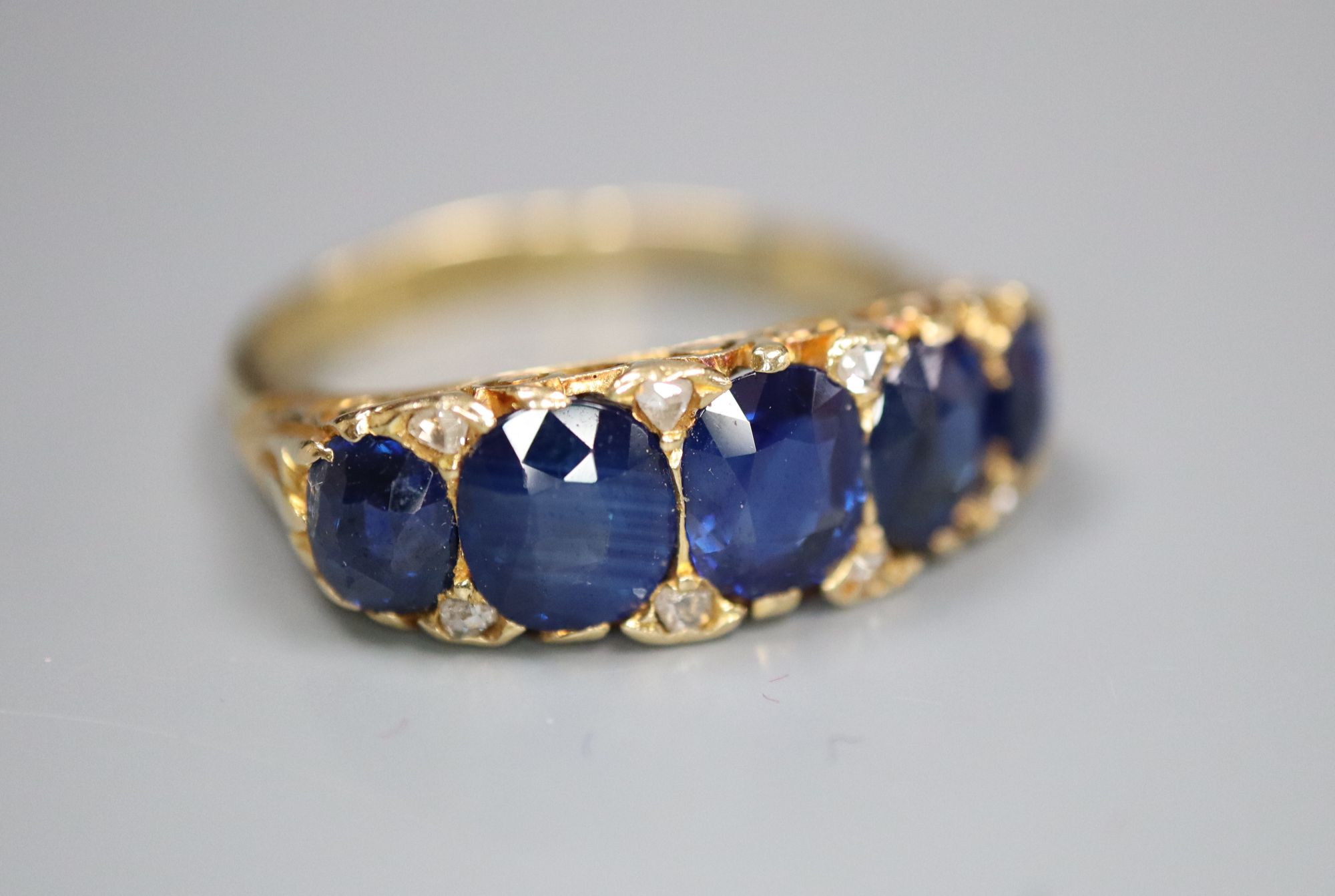 A George V 18ct gold and graduated five stone sapphire set half hoop ring, with diamond chip spacers,